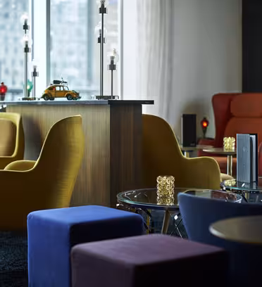 En modern loungedel med gula och röda fåtöljer, blå och lila kubstolar och små runda glasbord. En trädisk med dekorativa lampor och en leksaksbil finns i bakgrunden, med stora fönster med utsikt över staden.