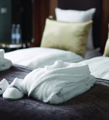 Ett mysigt hotellrum har en snyggt bäddad säng med två guldkuddar. På sängen ligger två fluffiga vita badrockar och ett par vita tofflor. I bakgrunden sitter två flaskvatten på ett sidobord.