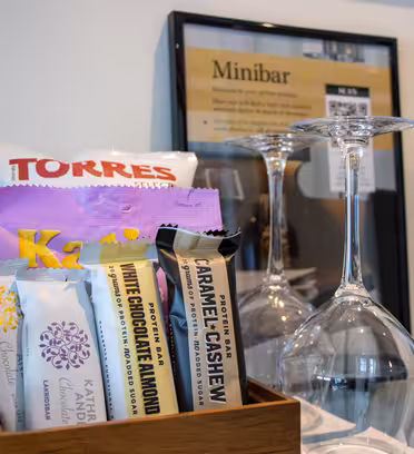 En minibar med en träbricka med snacks som chips, chokladkakor och nötter. Två vinglas står upp och ner bredvid en flaska rött vin. En inramad meny finns i bakgrunden.