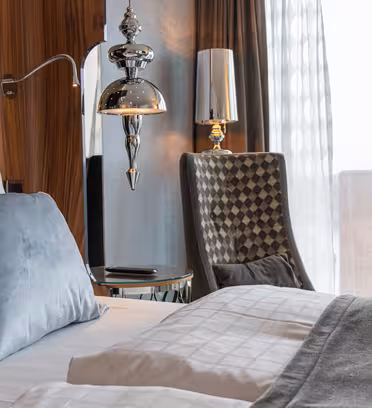 Hotellrum med en snyggt bäddad säng med vita sängkläder och en blå accentkudde. En modern lampa och stol sitter bredvid en väggpanel i trä. Ett fönster med skira gardiner erbjuder utsikt över en stadsbyggnad.