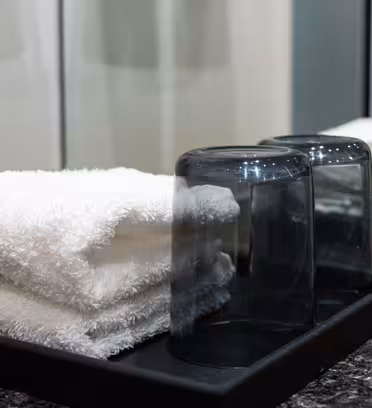 En snyggt arrangerad svart bricka rymmer två vita vikta handdukar och två upp och ner genomskinliga glas på en spräcklig bänkskiva, med en spegel som reflekterar scenen.