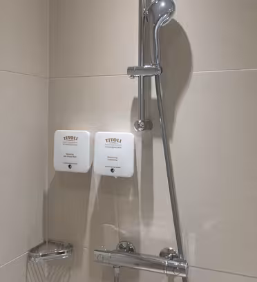 Duschområde med ett modernt duschhuvud i krom och två väggmonterade vita dispensrar märkta "Tivoli". Dispensrarna innehåller schampo och duschgel. Beige kakel täcker väggarna och en liten metallkorg är fäst under dispensrarna.