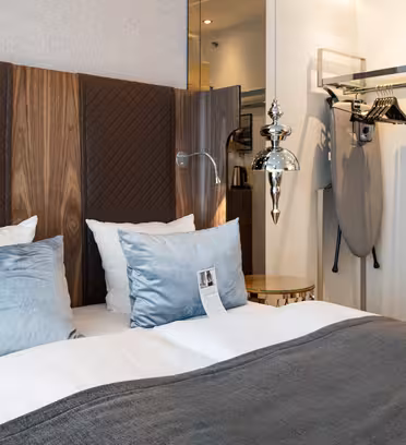 Ett prydligt arrangerat hotellrum med en säng med vita och gråa sängkläder, blågrå kuddar och en sänggavel i trä. På sidan syns ett strykjärn och strykbräda tillsammans med en vattenkokare på en hylla. En modern taklampa hänger ovanför.