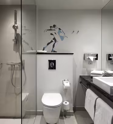 Modernt badrum med glasdusch, toalett och stor spegel. Väggarna är ljusgråa och på väggen finns dekorativ konst av en figur i rörelse. Handfatet har ett vitt handfat, toalettartiklar och snyggt vikta handdukar.