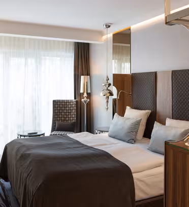 Ett modernt hotellrum med en snyggt bäddad dubbelsäng med mörka sängkläder, två stolar vid ett stort fönster med skira gardiner och ett litet skrivbord med en lampa. Rummet är inrett i neutrala toner med moderna belysningsarmaturer.