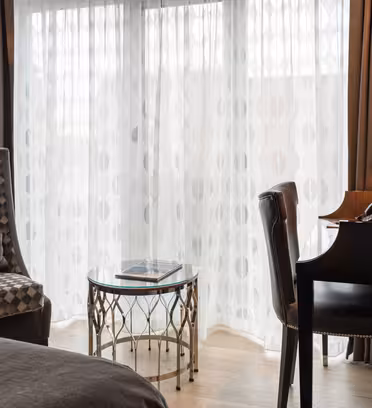 Ett modernt hotellrum har en rutig fåtölj, ett litet glasbord och ett mörkt skrivbord med en lampa. Skira gardiner hänger framför ett stort fönster och släpper in diffust naturligt ljus i rummet. En säng är delvis synlig.