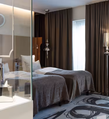 Ett modernt hotellrum har två enkelsängar med grå sängkläder, flankerad av en glasparti med ett vitt handfat och kranar. I taket hänger mörka gardiner och en mönstrad matta ger golvet en dekorativ touch.