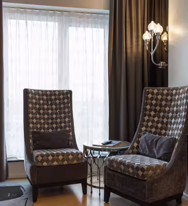 Ett modernt hotellrum har två högryggade stolar med geometriska mönster, ett litet bord med glasskiva och en väggmonterad TV. Gardiner från golv till tak täcker ett fönster, och en golvlampa står i närheten. En vägglampa ger extra ljus.