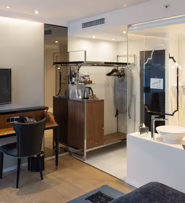 Modernt hotellrum med väggmonterad TV, skrivbord, stol och öppen garderob med hängare, värdeskåp och kaffebryggare. En glasvägg skiljer badrumsdelen åt med ett handfat och en stor spegel. Neutrala toner skapar en elegant, modern känsla.