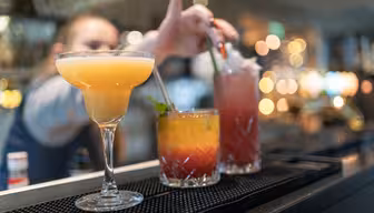 Närbild av tre färgglada cocktails uppradade på en bar. Den första är ljusgul i ett krökt glas, den andra är en lager orangeröd drink garnerad med mynta, och den tredje är en hög rosa dryck med sugrör. Suddig bartender i bakgrunden.