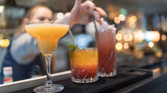 Närbild av tre färgglada cocktails uppradade på en bar. Den första är ljusgul i ett krökt glas, den andra är en lager orangeröd drink garnerad med mynta, och den tredje är en hög rosa dryck med sugrör. Suddig bartender i bakgrunden.
