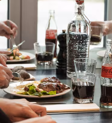 Människor som äter vid ett bord med tallrikar med mat, inklusive sallad och kött. Flaskor och glas Coca-Cola och andra drycker står på bordet. Händer ses använda redskap och hålla glasögon. Inställningen verkar avslappnad och mysig.