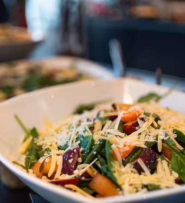 En närbild av en fräsch sallad med spenat, skivade persikor, plommon och riven ost i en vit skål. Bakgrunden visar suddiga rätter, vilket tyder på en buffémiljö.