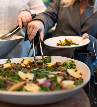 Två personer serverar sig sallad från stora skålar. Salladen innehåller grönsaker, äppelskivor och nötter. En person använder en tång för att placera sallad på en vit tallrik. En burk med frön och oljeflaskor står i närheten på bordet.