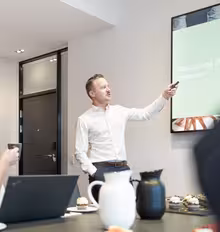 En man i en vit skjorta presenterar i ett konferensrum och pekar på en skärm som visar en bild. En kvinna sitter vid ett bord med en bärbar dator och en kopp och lyssnar. Rummet har en modern design med snacks synliga på bordet.