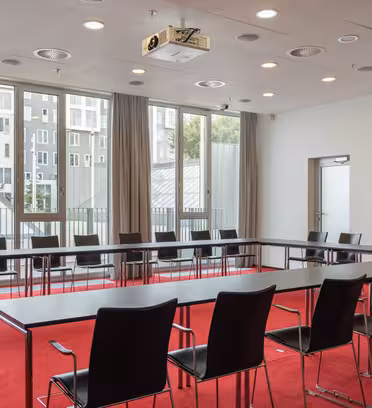 Ett modernt konferensrum med röd matta, stora fönster och ett U-format arrangemang av svarta stolar runt bord. Det finns en projektor i taket, en dörr till höger och en inramad bild på väggen. Naturligt ljus lyser upp rummet.