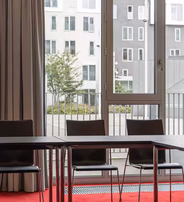 Ett modernt konferensrum med ett U-format arrangemang av svarta stolar och bord. Stora fönster med gardiner ger utsikt över moderna byggnader utanför. Golvet är täckt med en röd matta.