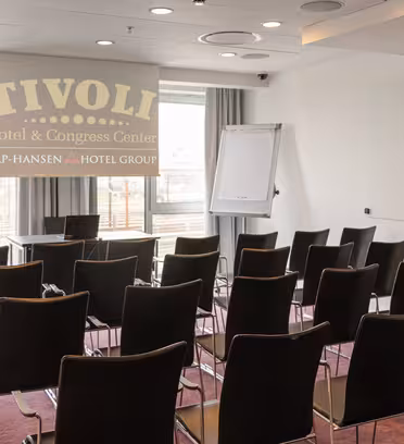 Litet konferensrum inrett med rader av tomma svarta stolar vända mot en projektorduk som visar "TIVOLI Hotel & Congress Center." En blädderblock och en whiteboard finns nära skärmen och stora fönster släpper in naturligt ljus utifrån.