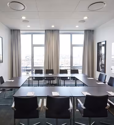 Ett modernt konferensrum med ett stort U-format bord, omgivet av nio svarta stolar. En platt-TV är monterad på vänster vägg, med en whiteboard till höger. Stora fönster släpper in naturligt ljus och gardiner är delvis fördragna.