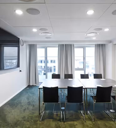 Ett modernt konferensrum med ett långbord omgivet av åtta stolar. En stor platt-TV är monterad på väggen till vänster. Rummet har fönster från golv till tak med gardiner och gröna heltäckningsmattor.