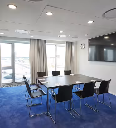 Ett modernt konferensrum med ett stort bord och åtta svarta stolar på en blå matta. En stor platt-TV är monterad på väggen, nära stora fönster med beiga gardiner. Rummet är väl upplyst och har utsikt över stadsbilden.