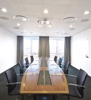 Ett modernt konferensrum med ett långt träbord omgivet av svarta stolar. Rummet har en whiteboard, en väggklocka och inramade konstverk på väggarna. Det finns stora fönster längst bort, så att naturligt ljus lyser upp utrymmet.