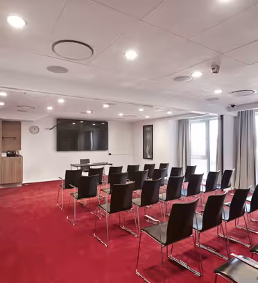 Ett modernt konferensrum har rader av svarta stolar placerade mot en vägg med en stor platt-TV. Rummet har röda mattor, vita väggar, ett träskåp och stora fönster med gardiner. Taklampor lyser upp utrymmet.