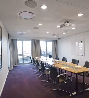 Ett modernt konferensrum med ett långt träbord och svarta stolar. Det finns en projektor i taket, en whiteboard på väggen, två stora fönster med gardiner och ett heltäckningsmatta.