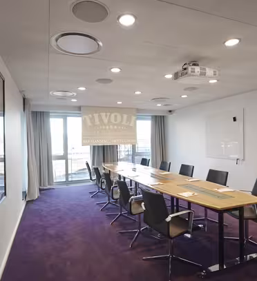 Ett modernt konferensrum med ett långt träbord och svarta stolar. Rummet har ett stort fönster med utsikt, whiteboard på väggen och takprojektorer. Golvet är täckt med lila matta.