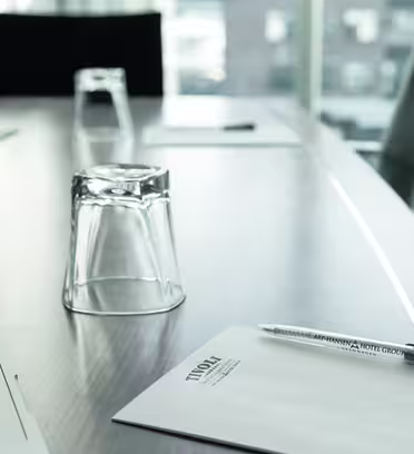 Ett konferensbord med glasögon, anteckningsblock och pennor snyggt arrangerat. Solljus filtreras genom stora fönster och lyser upp scenen. Två stolar är delvis synliga, vilket tyder på en professionell mötesinställning.