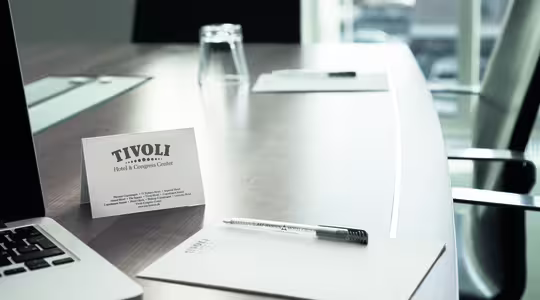 Ett mötesbord med en bärbar dator, två stolar, tomma glas och skrivblock. Fokus ligger på ett visitkort med texten "Tivoli Hotel & Congress Center", tillsammans med penna och papper, som föreslår en formell miljö för en konferens eller ett möte.