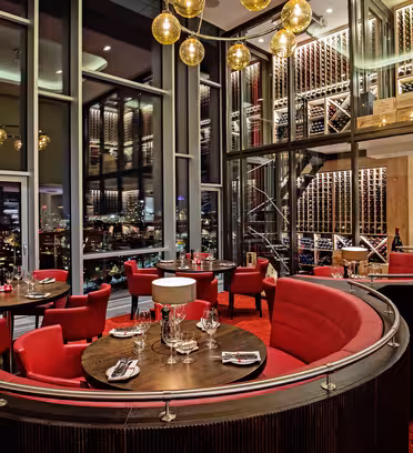 Lyxig restauranginteriör med runda bord för middagar, omgivna av eleganta röda stolar och svängda bås. Fönster från golv till tak avslöjar en stadsbild, och en framträdande vägg visar vinflaskor. Varm belysning förhöjer stämningen.