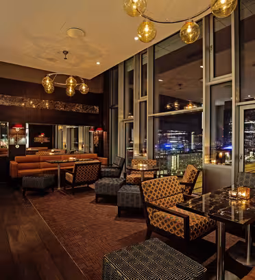 Elegant lounge med vadderade stolar och soffor, varm belysning från hängande klotlampor och bord med ljus. Fönster från golv till tak erbjuder en nattlig stadsutsikt, vilket skapar en mysig och sofistikerad atmosfär.