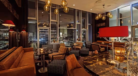 En mysig, sofistikerad lounge med bruna skinnsoffor och mönstrade stolar. Ett marmorbord pryds av dekorativa föremål. Stora fönster erbjuder utsikt över stadsbilden på natten, och varm belysning skapar en inbjudande atmosfär.