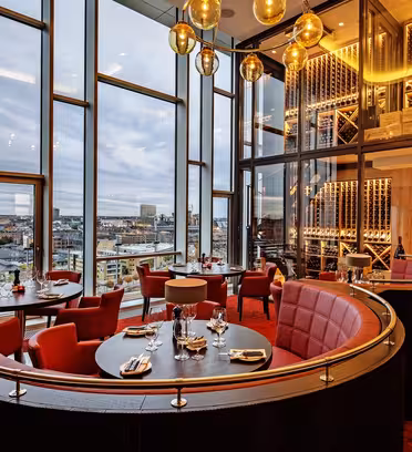 Lyxig restauranginteriör med röda sittplatser och elegant dukning. Stora fönster visar en stadssilhuett. Varm belysning från hängande jordglobsarmaturer och hyllor med vin bidrar till den sofistikerade atmosfären.