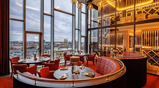 Lyxig restauranginteriör med röda sittplatser och elegant dukning. Stora fönster visar en stadssilhuett. Varm belysning från hängande jordglobsarmaturer och hyllor med vin bidrar till den sofistikerade atmosfären.