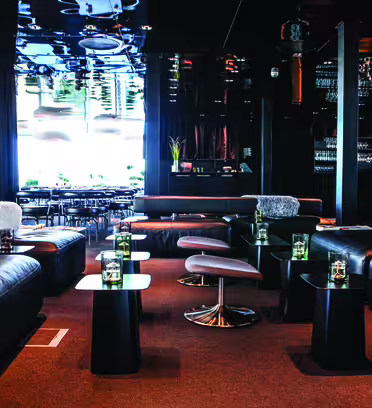En modern lounge med sittplatser i svart läder och snygga låga bord. Utrymmet har ett glänsande svart tak, svag belysning och ett barområde i bakgrunden. Ljusstakar i glas pryder varje bord och ger en touch av elegans.