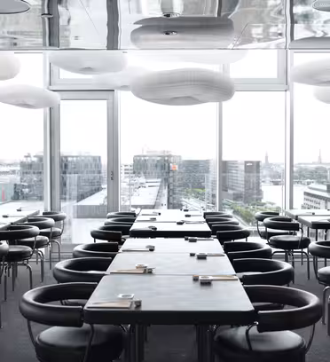 Modern, minimalistisk restaurang med rektangulära bord, svarta vadderade stolar och stora fönster med utsikt över stadsbilden. Vita, molnliknande taklampor skapar en mjuk, elegant atmosfär.