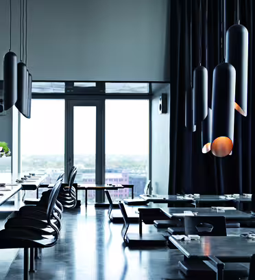 En modern, minimalistisk restauranginteriör med eleganta svarta bord och stolar. Stora taklampor hänger i taket och ett panoramafönster erbjuder utsikt utanför. Golvet är blankt och reflekterar det omgivande ljuset.