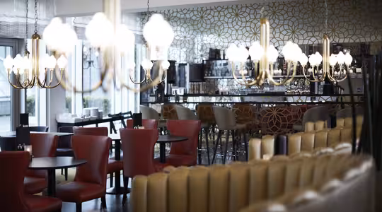 En modern restauranginredning med röda och beige stolar, mörka runda bord och eleganta ljuskronor. Bardelen med barnstolar syns i bakgrunden, mot en mönstrad vägg. Atmosfären är mysig och inbjudande.