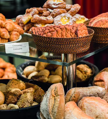 En visning av olika bakverk, inklusive korgar med croissanter, bakverk och skivat bröd. Det finns också olika frallor och bröd, som baguetter och rustikt bröd, på svarta och vävda korgar. En skylt märkt "croissanter" är synlig.
