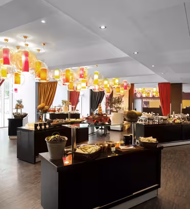 En rymlig bufférestaurang med elegant inredning. Borden är fyllda med en mängd olika matvaror och korgar, uppställda under färgglada taklampor. Stora fönster och livfulla gardiner ger ljusstyrka och färg till rummet.