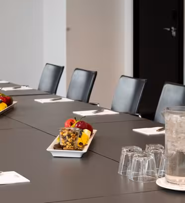 Ett modernt konferensrum med ett långbord omgivet av svarta stolar. Bordet är dukat med anteckningsböcker, pennor, en kanna vatten och glas. Pläterade snacks med frukt och nötter placeras i mitten. Rummet är väl upplyst och rent.