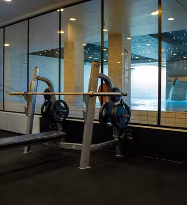 Ett gym med en bänkpress i förgrunden, med flera viktskivor på en skivstång. Stora fönster visar utsikten över en pool med forsande vatten, och området är väl upplyst med taklampor.