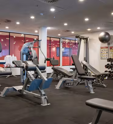 Ett modernt gym med olika träningsmaskiner, inklusive viktbänkar, en elliptisk träningsmaskin och en roddmaskin. Hantlar linjeställ i hörnet. Rummet är väl upplyst med taklampor och stora fönster reflekterar interiören.