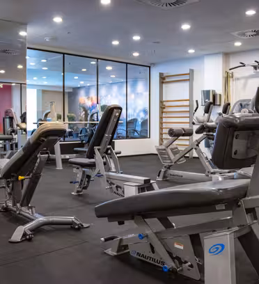 Ett modernt gym har olika träningsredskap, inklusive viktmaskiner och bänkar, placerade på ett mörkt golv. Speglar och stark belysning förstärker utrymmet, med färgglad väggkonst synlig genom en glasvägg i bakgrunden.