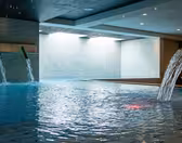 Inomhuspool med forsande vattenfunktioner och modern design. Poolen reflekterar takbelysning, med vatten som rinner från väggmonterade piper till det blåkaklade området, omgivet av ljusa väggar och tak.