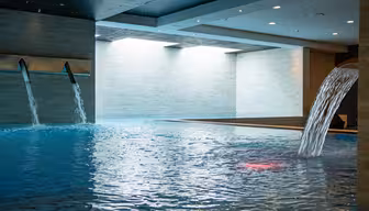 Inomhuspool med forsande vattenfunktioner och modern design. Poolen reflekterar takbelysning, med vatten som rinner från väggmonterade piper till det blåkaklade området, omgivet av ljusa väggar och tak.