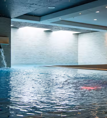 Inomhuspool med forsande vattenfunktioner och modern design. Poolen reflekterar takbelysning, med vatten som rinner från väggmonterade piper till det blåkaklade området, omgivet av ljusa väggar och tak.