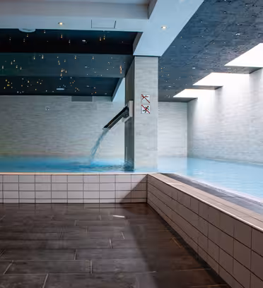 Inomhuspool med modern design, med en vattenkaskad från en kolumn. Poolen är omgiven av ljust kakel och taket har stjärnliknande ljus. Takfönster tillåter naturligt ljus att lysa upp området.